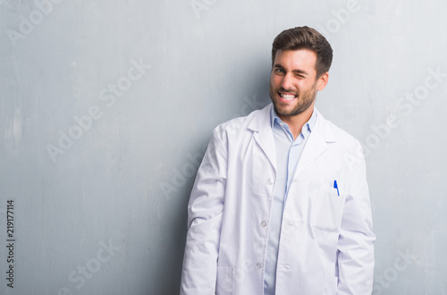 Handsome young professional man over grey grunge wall wearing white coat winking looking at the camera with sexy expression, cheerful and happy face.