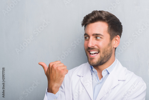 Handsome young professional man over grey grunge wall wearing white coat smiling with happy face looking and pointing to the side with thumb up.