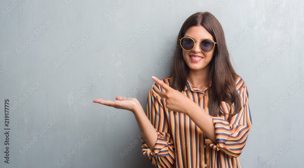 Young brunette woman over grunge grey wall wearing retro sunglasses and outfit very happy pointing with hand and finger