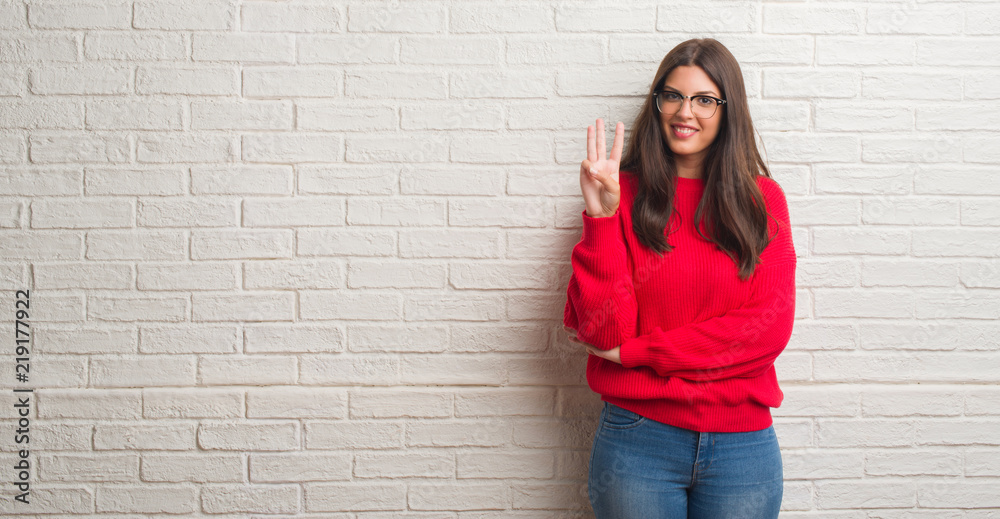 Young brunette woman standing over white brick wall showing and pointing up with fingers number three while smiling confident and happy. Young brunette woman standing over white brick wall showing and pointing up with fingers number three while smiling confident and happy.