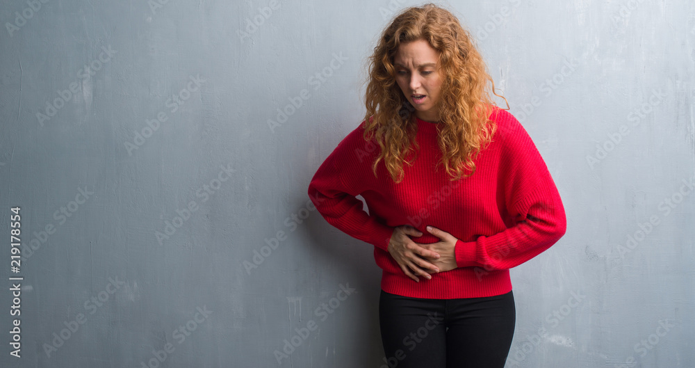 Young redhead woman over grey grunge wall wearing red sweater with hand on stomach because indigestion, painful illness feeling unwell. Ache concept. Young redhead woman over grey grunge wall wearing red sweater with hand on stomach because indigestion, painful illness feeling unwell. Ache concept.