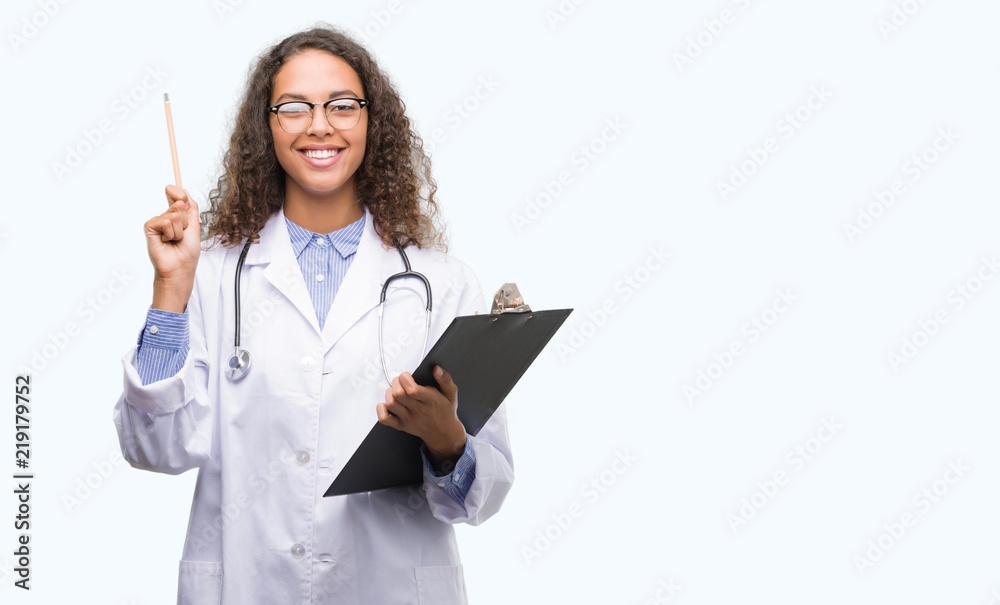 Young hispanic doctor woman holding a clipboard surprised with an idea or question pointing finger with happy face, number one Young hispanic doctor woman holding a clipboard surprised with an idea or question pointing finger with happy face, number one