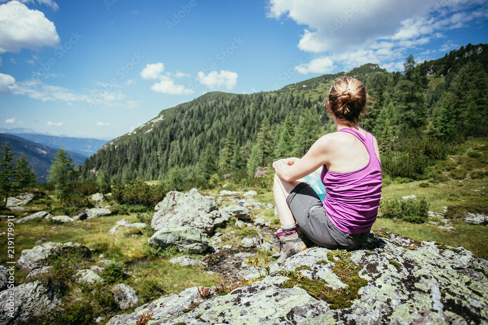 Junge Frau in Wanderschuhen sitzt auf Felsbrocken und genießt die Aussicht über die Berglandschaft