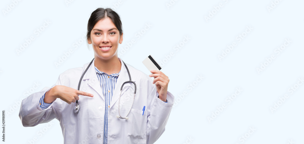 Young hispanic doctor woman holding credit card with surprise face pointing finger to himself Young hispanic doctor woman holding credit card with surprise face pointing finger to himself