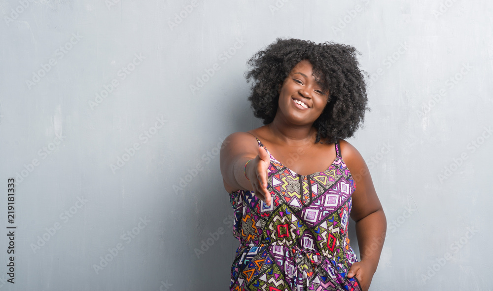 Young african american woman over grey grunge wall wearing colorful dress smiling friendly offering handshake as greeting and welcoming. Successful business.