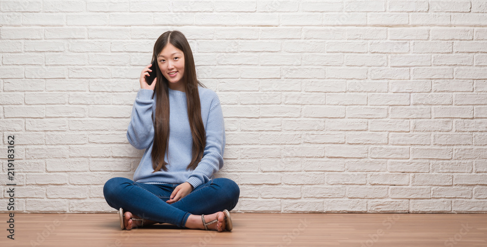 Young Chinese woman sitting on the floor over brick wall with a happy face standing and smiling with a confident smile showing teeth Young Chinese woman sitting on the floor over brick wall with a happy face standing and smiling with a confident smile showing teeth