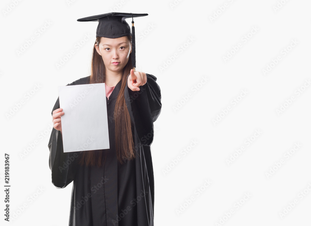 Young Chinese woman wearing graduate uniform holding paper degree pointing with finger to the camera and to you, hand sign, positive and confident gesture from the front Young Chinese woman wearing graduate uniform holding paper degree pointing with finger to the camera and to you, hand sign, positive and confident gesture from the front