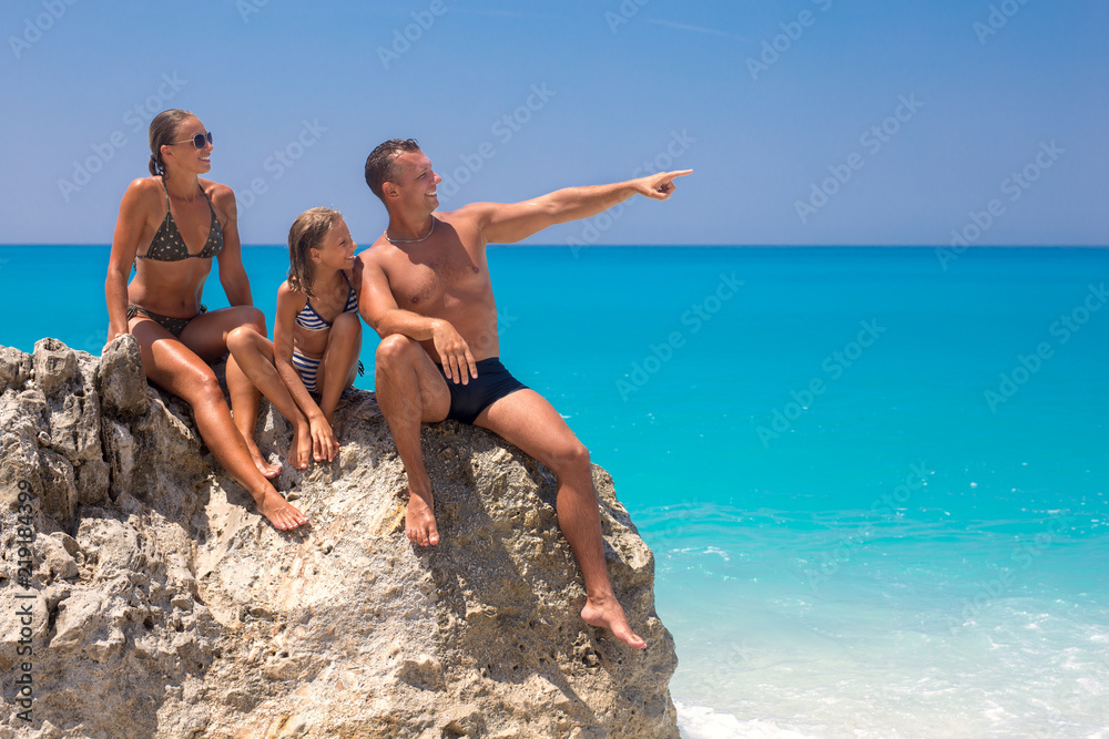 Fototapeta premium Happy young family sitting on a rock at the beach looking at something