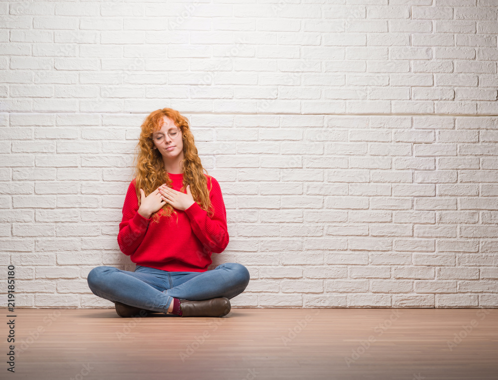 Young redhead woman sitting over brick wall smiling with hands on chest with closed eyes and grateful gesture on face. Health concept. Young redhead woman sitting over brick wall smiling with hands on chest with closed eyes and grateful gesture on face. Health concept.