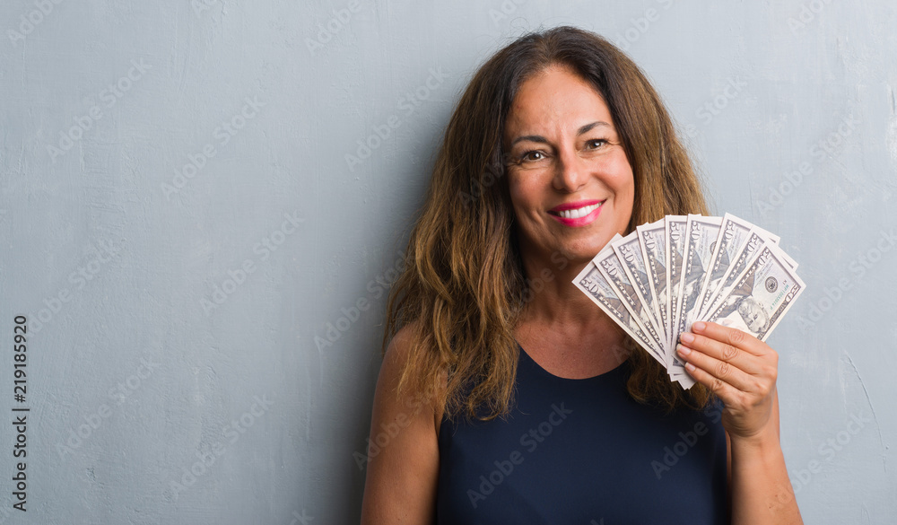 Middle age hispanic woman standing over grey grunge wall holding dollars with a happy face standing and smiling with a confident smile showing teeth Middle age hispanic woman standing over grey grunge wall holding dollars with a happy face standing and smiling with a confident smile showing teeth