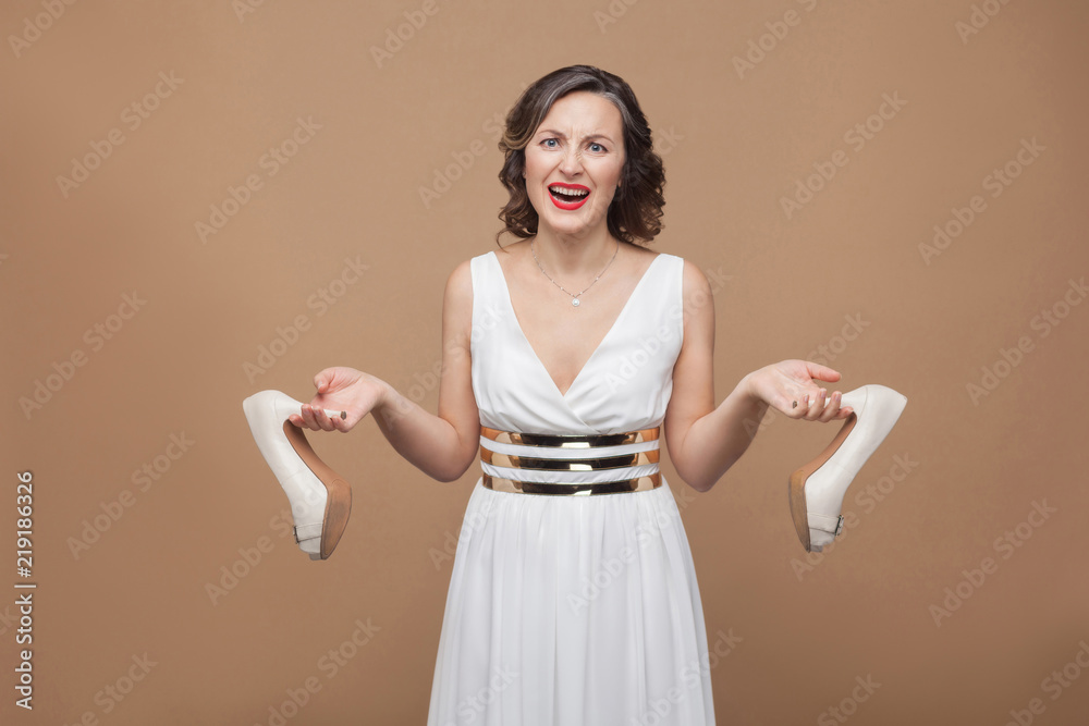 Angry business woman in white dress standing looking at camera screaming and holding uncomfortable high heels. Emotion and feeling concept. Studio shot, indoor, isolated on light brown background Angry business woman in white dress standing looking at camera screaming and holding uncomfortable high heels. Emotion and feeling concept. Studio shot, indoor, isolated on light brown background