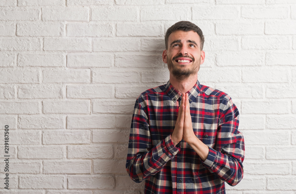 Young adult man standing over white brick wall begging and praying with hands together with hope expression on face very emotional and worried. Asking for forgiveness. Religion concept. Young adult man standing over white brick wall begging and praying with hands together with hope expression on face very emotional and worried. Asking for forgiveness. Religion concept.