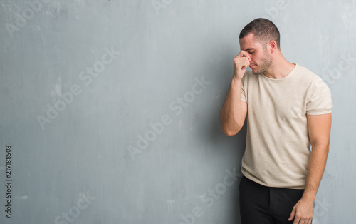 Young caucasian man over grey grunge wall tired rubbing nose and eyes feeling fatigue and headache. Stress and frustration concept.