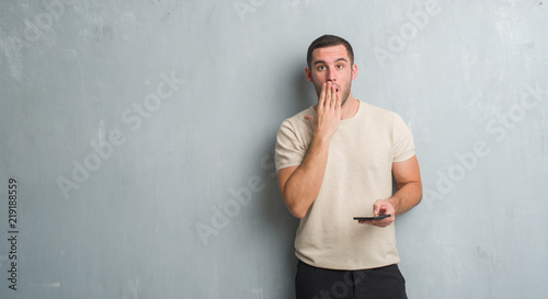 Young caucasian man over grey grunge wall texting a message using smartphone cover mouth with hand shocked with shame for mistake, expression of fear, scared in silence, secret concept