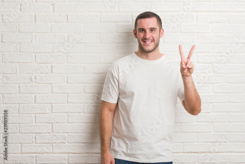 Young caucasian man standing over white brick wall smiling with happy face winking at the camera doing victory sign. Number two.