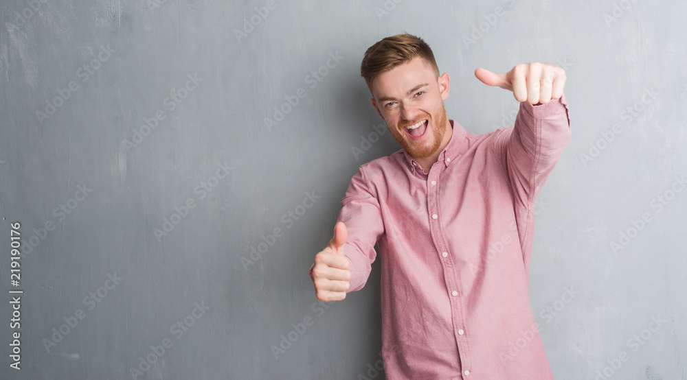 Young redhead man over grey grunge wall wearing pink shirt approving doing positive gesture with hand, thumbs up smiling and happy for success. Looking at the camera, winner gesture. Young redhead man over grey grunge wall wearing pink shirt approving doing positive gesture with hand, thumbs up smiling and happy for success. Looking at the camera, winner gesture.
