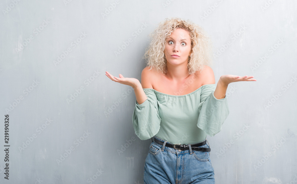 Young blonde woman with curly hair over grunge grey background clueless and confused expression with arms and hands raised. Doubt concept. Young blonde woman with curly hair over grunge grey background clueless and confused expression with arms and hands raised. Doubt concept.