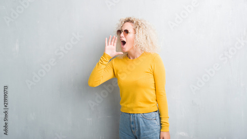 Young blonde woman with curly hair over grunge grey background shouting and screaming loud to side with hand on mouth. Communication concept.