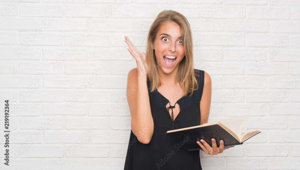 Beautiful young woman over white brick wall reading a book very happy and excited, winner expression celebrating victory screaming with big smile and raised hands Beautiful young woman over white brick wall reading a book very happy and excited, winner expression celebrating victory screaming with big smile and raised hands