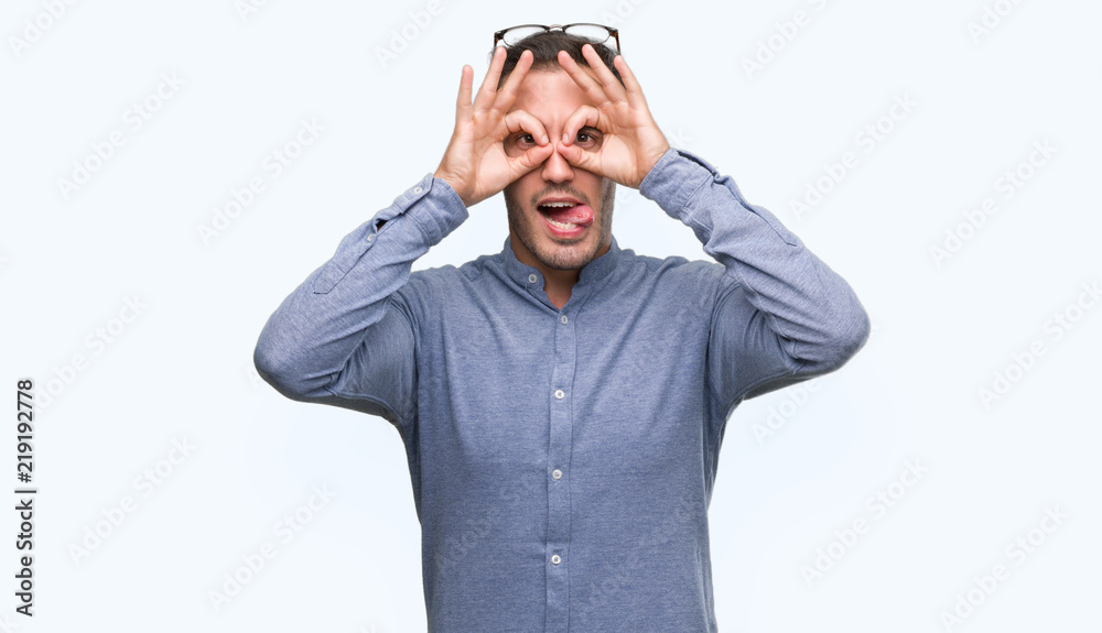 Handsome young elegant man wearing glasses doing ok gesture like binoculars sticking tongue out, eyes looking through fingers. Crazy expression. Handsome young elegant man wearing glasses doing ok gesture like binoculars sticking tongue out, eyes looking through fingers. Crazy expression.