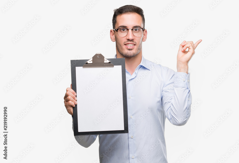 Handsome young business man showing a clipboard very happy pointing with hand and finger to the side Handsome young business man showing a clipboard very happy pointing with hand and finger to the side
