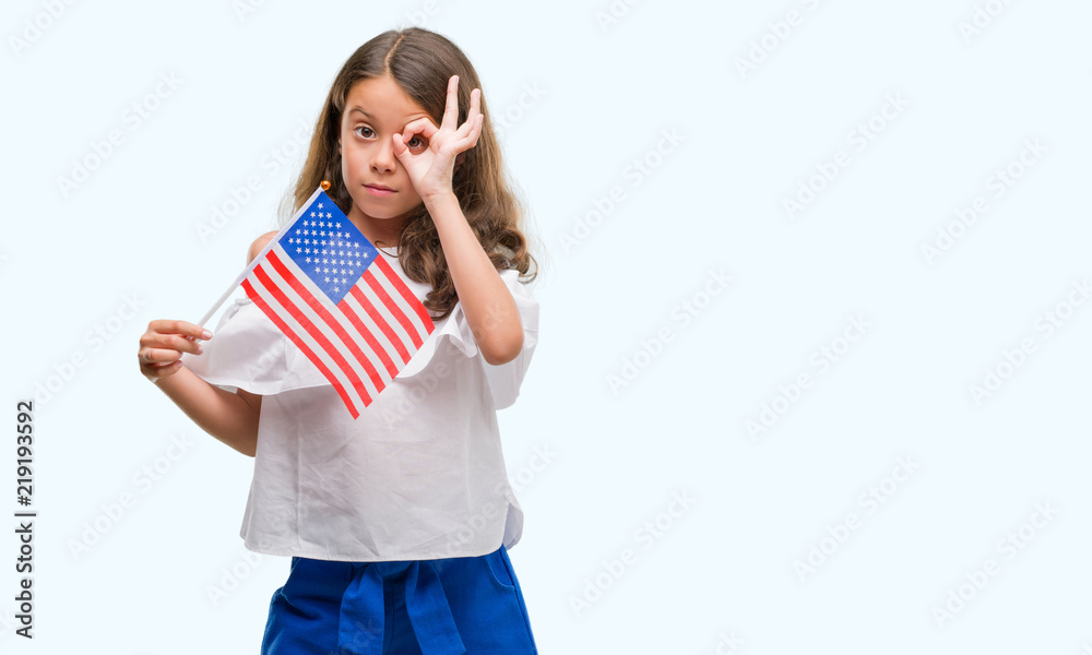 Brunette hispanic girl holding flag of United States of America with happy face smiling doing ok sign with hand on eye looking through fingers Brunette hispanic girl holding flag of United States of America with happy face smiling doing ok sign with hand on eye looking through fingers