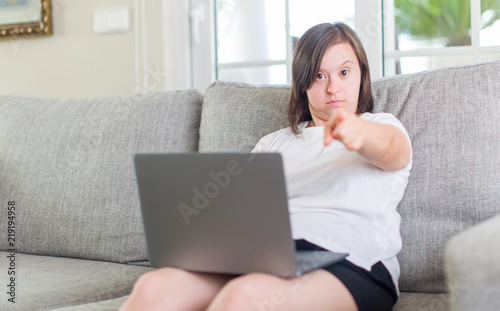 Down syndrome woman at home using computer laptop pointing with finger to the camera and to you, hand sign, positive and confident gesture from the front