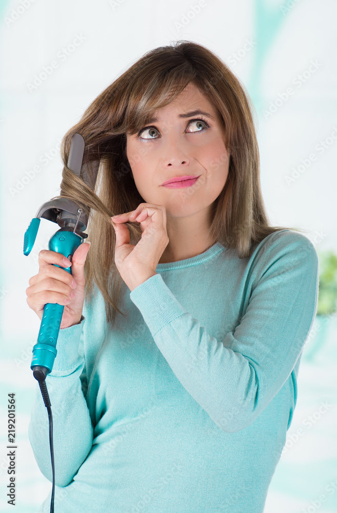 Close up of a beautiful young woman using a hair straighteners to do curly hair by herslef, in a blurred background Close up of a beautiful young woman using a hair straighteners to do curly hair by herslef, in a blurred background