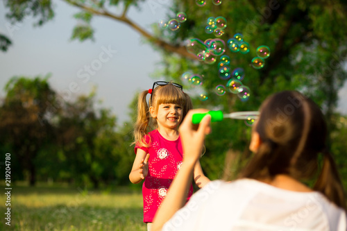 Young mother and little daughter playing in park with soap bubbles. Love family, parenthood, childhood