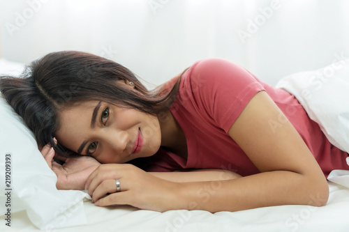 Attractive cheerful young woman lying down with pillow after sleeping on white bed looking at camera.