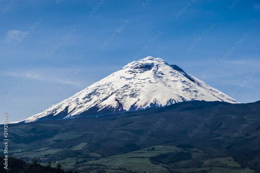 Frontal view of the Cotopaxi volcano in Ecuador. It's a sunny day with almost no clouds and blue sky. Trees and agriculture land can be seen in the front.