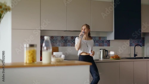 Athletic girl in t-shirt and fitness leggings drinking water from bottle. Young woman in fashion sportswear relaxing in kitchen. Beautiful girl listening music in headphones. After morning run