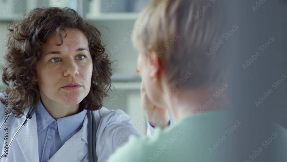 Beautiful female doctor using penlight for eye exam of female patient in clinic and then smiling and speaking while writing prescription Beautiful female doctor using penlight for eye exam of female patient in clinic and then smiling and speaking while writing prescription