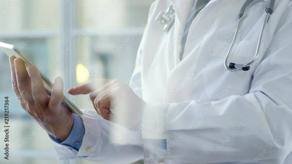 Close-up of unrecognizable male doctor in lab coat with stethoscope over neck standing in office and using digital tablet