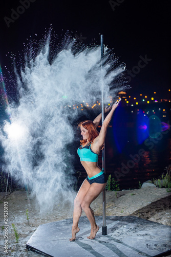 The red-haired woman in shorts and a top, dances on a pole and throws a flour against city line of a city. A young woman dances a striptease and throws up her hands with a white powder