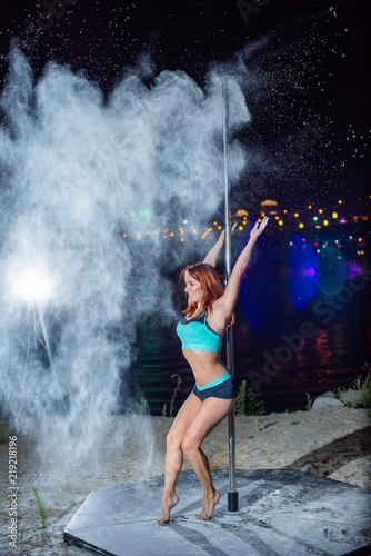 The red-haired woman in shorts and a top, dances on a pole and throws a flour against city line of a city. A young woman dances a striptease and throws up her hands with a white powder