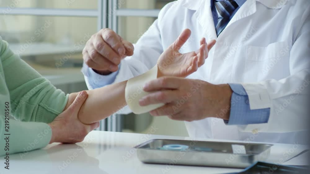 Tilt up of senior male doctor unwrapping elastic bandage from wrist of female patient and telling her to move fingers after recovery Tilt up of senior male doctor unwrapping elastic bandage from wrist of female patient and telling her to move fingers after recovery