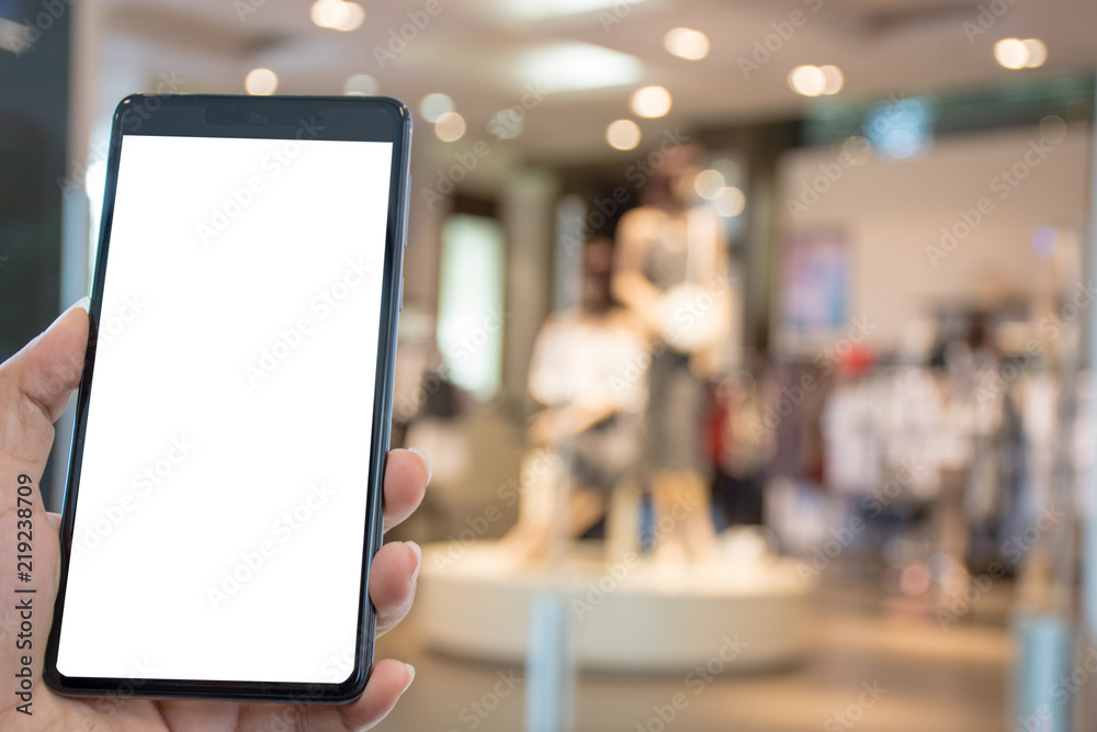 Fototapeta premium Female hand holding and showing mobile smartphone with blank white screen with boutique shop and model blurred background.