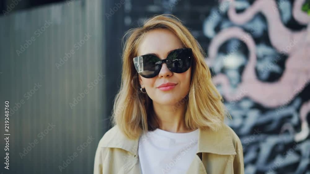 Portrait of beautiful blond girl in sunglasses looking at camera, laughing and touching her hair standing outdoors. Youth, fashion and emotions concept. Portrait of beautiful blond girl in sunglasses looking at camera, laughing and touching her hair standing outdoors. Youth, fashion and emotions concept.