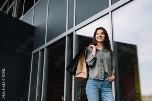 Successful positive woman with beautiful smile walking on street with shopping bags near modern supermarket or shopping center. Stylish caucasian female waiting taxi outdoors. 