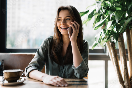 Young beautiful woman using mobile phone in modern coffee shop. Cute smiling businesswoman holding smartphone, sitting at working place. 