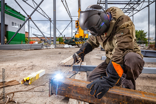A strong man is a welder in brown uniform, welding mask and welders leathers, a metal product is welded with a arc welding machine at the construction site, blue sparks fly to the sides photo