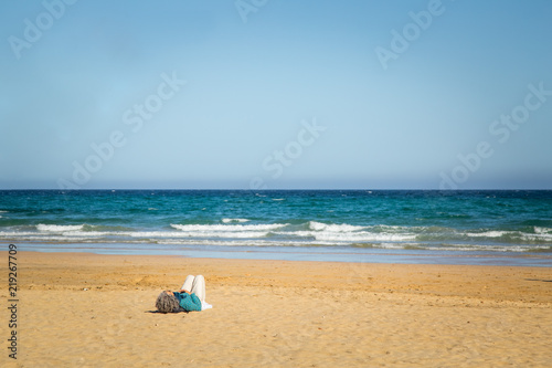 Elderly woman lying down on sandy beach with calm water in Fuerteventura.