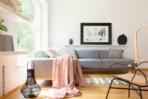 Pastel pink blanket thrown on corner couch standing in white living room interior with simple poster, lantern, decor and big window in the real photo