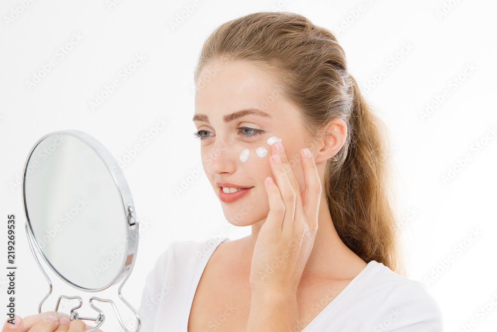 Young caucasian blonde woman with mirror and face skin care moisturizing cream on face isolated on white background. Anti aging and spa concept. Make up and nature beauty. Copy space Young caucasian blonde woman with mirror and face skin care moisturizing cream on face isolated on white background. Anti aging and spa concept. Make up and nature beauty. Copy space