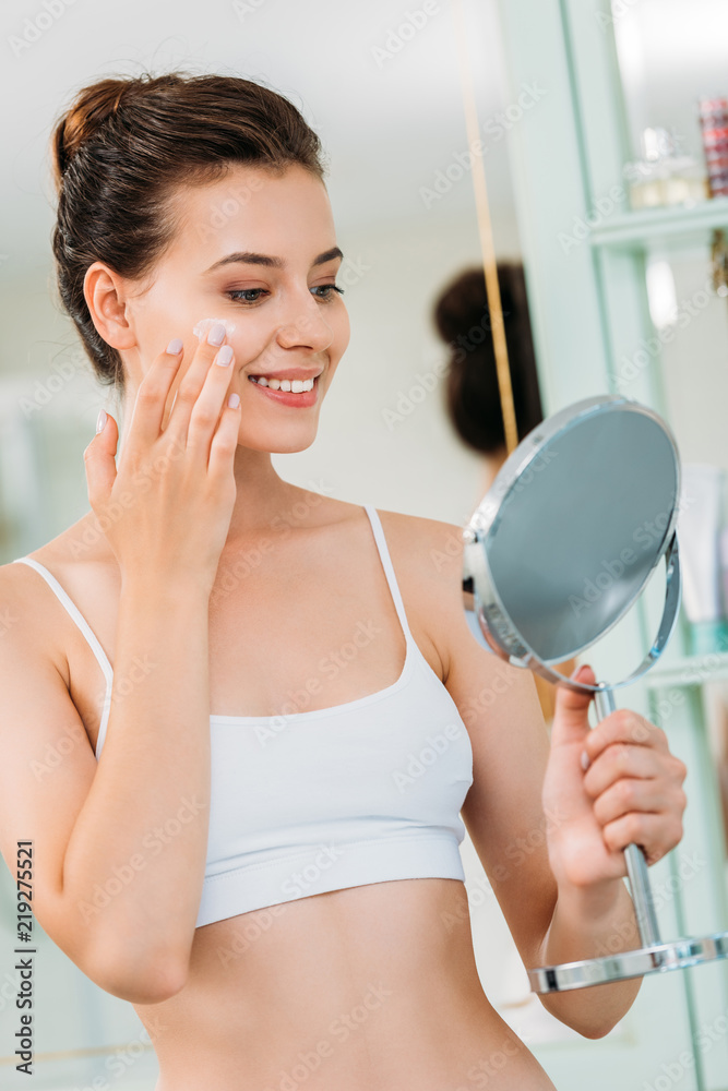 smiling young woman holding mirror and applying facial cream in bathroom smiling young woman holding mirror and applying facial cream in bathroom