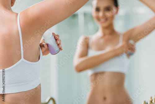 selective focus of smiling girl applying roller deodorant at mirror in bathroom