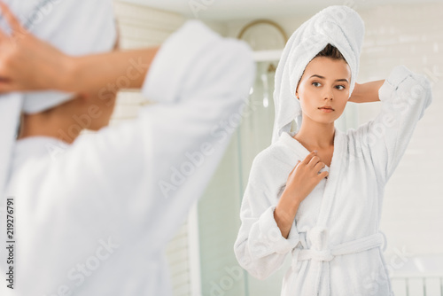beautiful young woman in bathrobe and towel on head looking at mirror in bathroom