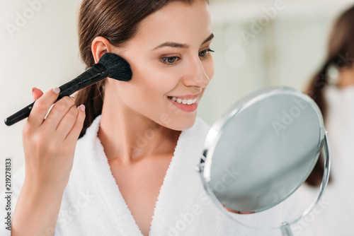 beautiful smiling brunette girl in bathrobe applying powder bronzer with brush photo