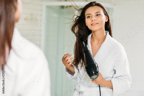beautiful smiling girl in bathrobe using hair dryer in bathroom